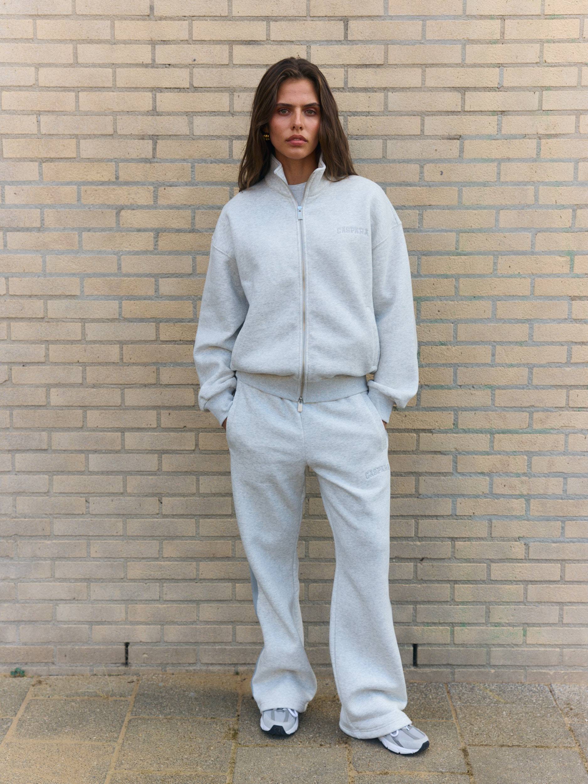 Woman wearing a grey melange funnel zip jacket and matching sweatpants against a brick wall.